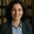Portrait of a professional woman with dark curly hair, smiling confidently indoors with a bookshelf background, representing expertise in history and storytelling.